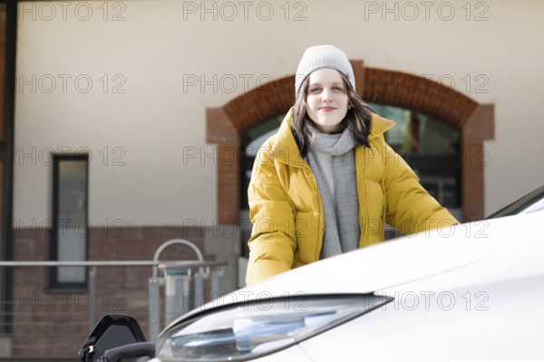Woman in yellow jacket smiles friendly, at charging station with electric car, environmentally friendly