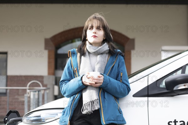 Woman with eyes closed holding a cup and leaning against a charging electric car, environmentally friendly