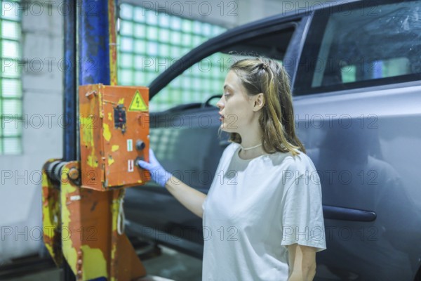 A woman in a workshop thoughtfully examines a switch on a car lift, partially obscured by the car