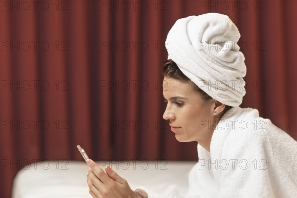 Young woman in bathrobe lying in hotel room using mobile phone, relaxed after taking a bath