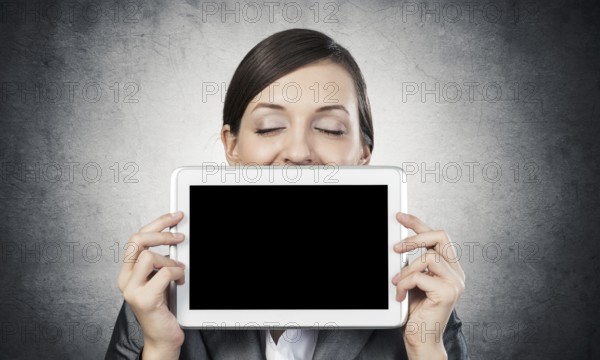 Businesswoman holding tablet computer with blank screen. Beautiful woman with closed eyes showing tablet PC near her face. Corporate businessperson on grey wall background. Digital technology