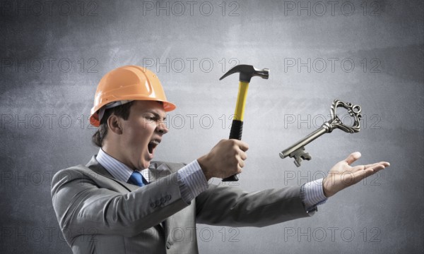 Furious businessman going to crash with hammer steel key. Young handsome contractor in business suit and safety helmet standing on grey wall background. Financial crisis in real estate. Password hack