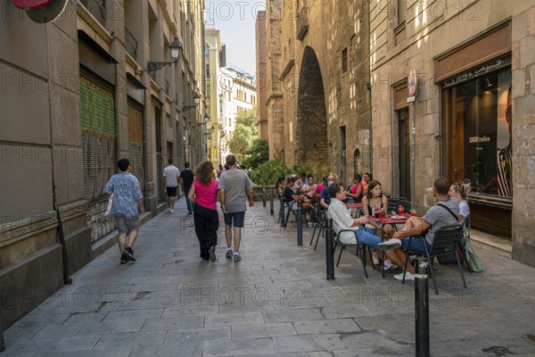 08-28-2022 Barcelona, SP People walking and sitting in cafe of Barcelona at narrow street in shadow (hot summer in stone capital city of Catalounia, Spain)
