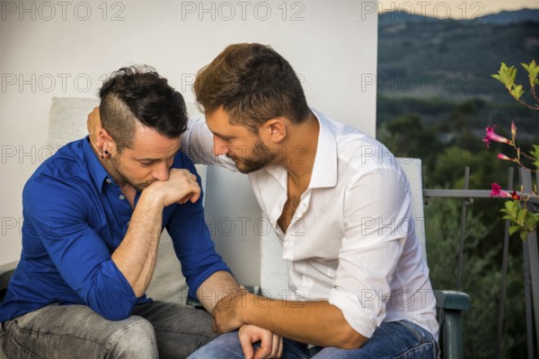 Portrait of two handsome men having trouble in relationship while sitting hugging and sad on balcony chairs