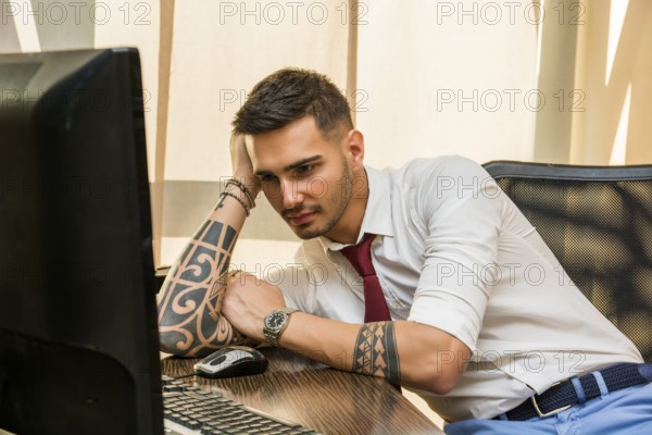 Tired bored young businessman sitting at his desk in front of his computer yawning, holding his head with his hand and eyes closed, in his office