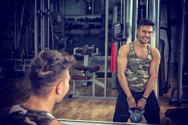 Attractive young athletic man exercising in gym, working out using kettlebell, looking at camera with a smile