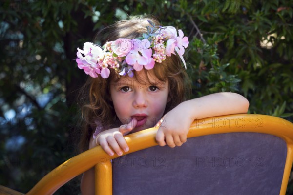 Happy childhood concept. Charming curly little girl in a wreath of flowers resting in a chair in the summer garden