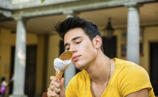 Funny attractive young man sitting, wearing yellow T-shirt while eating a tasty vanilla ice cream outdoors in the park, in a warm day of summer