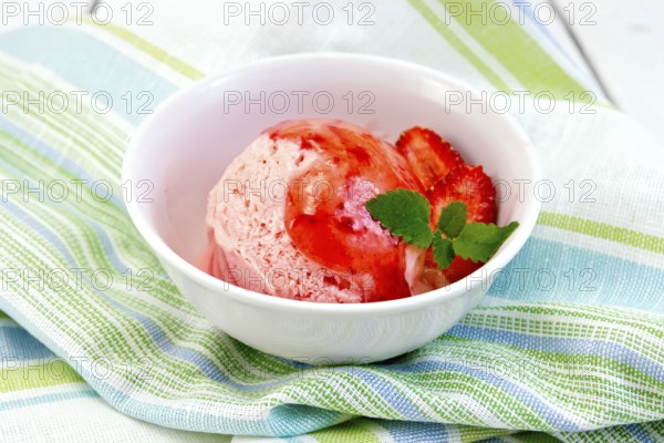Strawberry ice cream in a white bowl with strawberries and strawberry syrup on a striped linen napkin on a wooden boards background