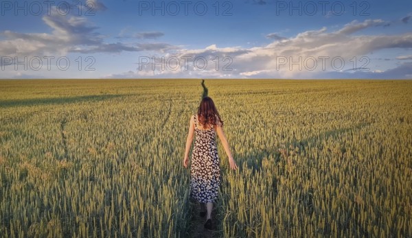 Carefree woman walking a pathway across a green wheat field. Idyllic countryside summer scenery, freedom concept