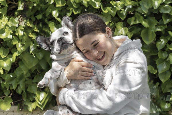 A girl in a white sweatshirt smiling and hugging a French Bulldog in a garden with lush green leaves in the background