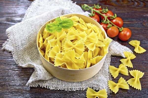 Farfalle pasta with basil in a bark box on a burlap napkin, tomatoes against dark wooden board
