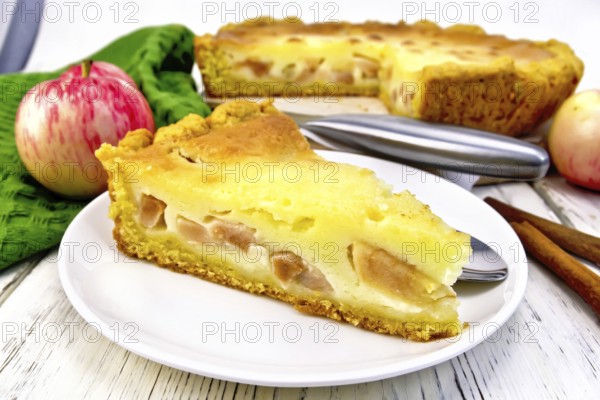 Piece of apple pie with sour cream sauce in a white plate, cinnamon, fruit and napkin on a wooden board background