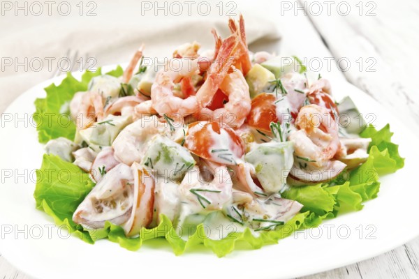 Salad with shrimp, avocado, tomatoes and mayonnaise on the lettuce in the plate, napkin, fork on background wooden plank