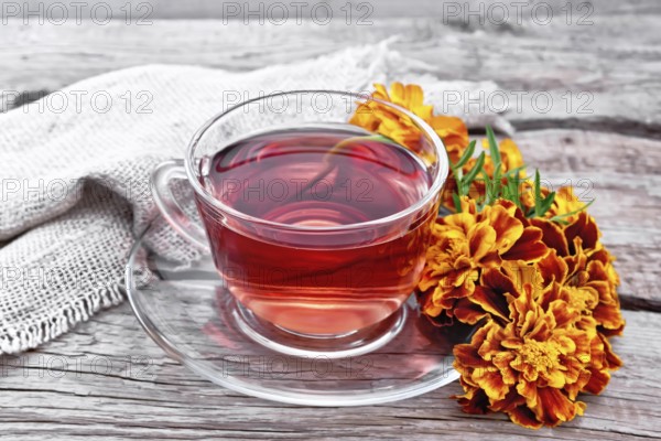 Marigold herbal tea in a glass cup and saucer, fresh flowers, sackcloth napkin on wooden board background