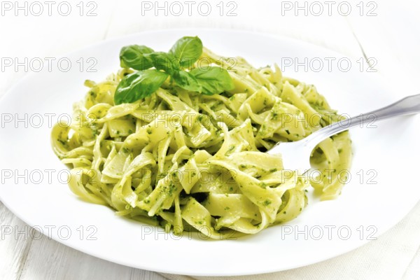 Tagliatelle pasta with pesto, basil and fork in a plate on a napkin against the background of light wooden board