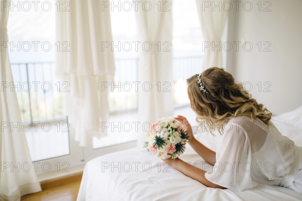 A bride in a gentle peignoir with a bouquet in her hands laying on the bed by the window with white curtains.