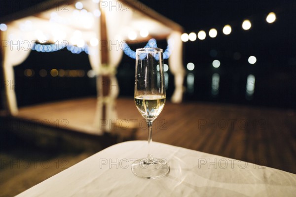 A glass of champagne stands on the table against the backdrop of a party on the beach. Beach talk with garlands at night.