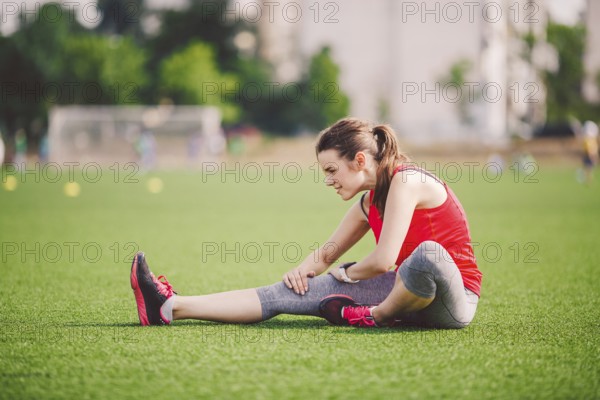 Theme sport and health. Young beautiful Caucasian woman sitting doing warm-up, warming up muscles, stretching green graß Restoration and rest of girl on stadium in summer on artificial turf