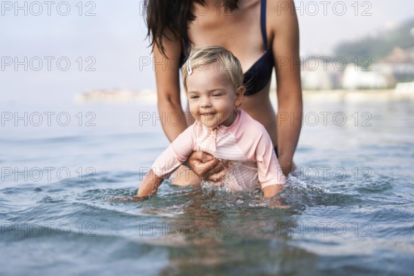 Little girl bathes in shallow water supported by her mom.