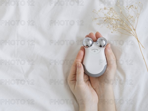 Microcurrent concept. Face massager with micro currents for home use and skincare on white bed. Female hands holds device for anti-aging and face lifting. Top view or flat lay, copy space for text