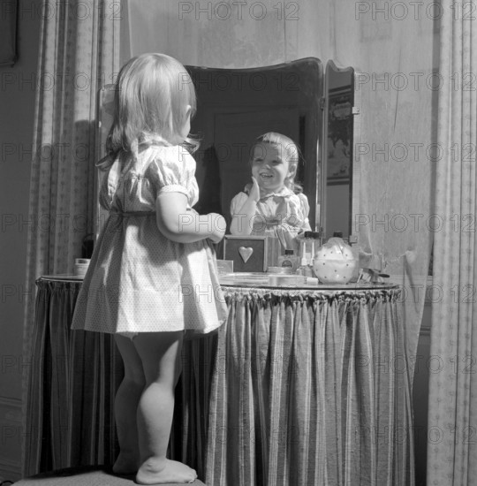 In the 1950s. A little girl is standing in front of her mothers makeup table and mirror and puts on some kind of beauty cream on her face, looking happy. Sweden 1951 Conard ref 1708
