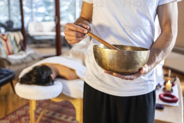 Massage therapist standing with a singing bowl in hands and giving a sound massage, to his client lying on the massage bed at the background