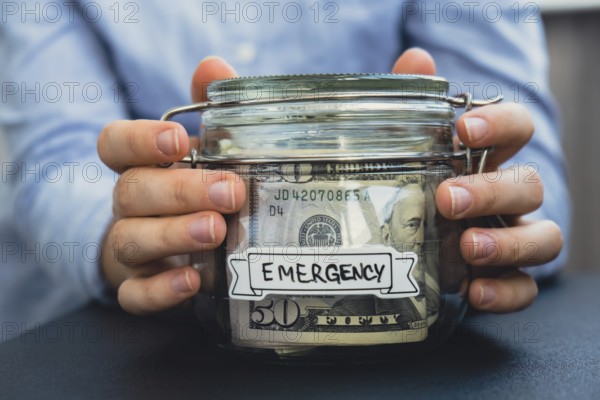 Female hands Saving Money In Glass Jar filled with Dollars banknotes. EMERGENCY transcription in front of jar. Managing personal finances extra income for future insecurity background