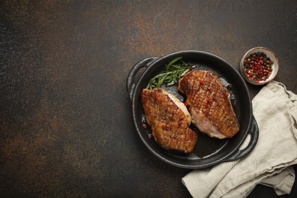 Two roasted duck breast fillets with crispy skin, with pepper and rosemary, top view in black cast iron pan with knife, dark brown concrete rustic background, space for text, food photography, food photography