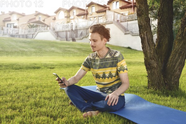 Young stylish handsome guy is sitting on grass and talking on the phone resting in the park. Taking a Break