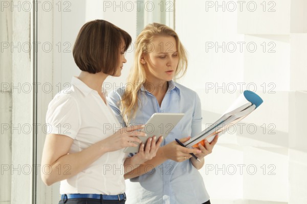 Two businesswomen standing at window and talking while holding tablet and notebook