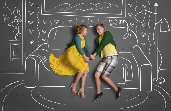 Happy valentines love story concept of a romantic couple sitting on a sofa and holding hands against chalk drawings background of a living room