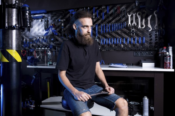 Portrait of small business owner of young man with beard. Guy bicycle mechanic workshop worker sitting with tool in his hand in a working black clothes in an apron in the background of a bicycle shop