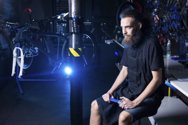 Portrait of small business owner of young man with beard. Guy bicycle mechanic workshop worker sitting with tool in his hand in a working black clothes in an apron in the background of a bicycle shop
