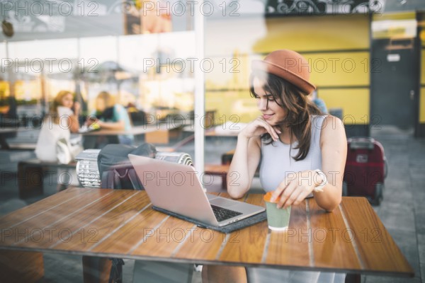 Beautiful young caucasian woman sitting on a terrace in a cafe in summer at a wooden table in a hat and a plate uses technology, working behind a laptop booking hotels trip in the summer