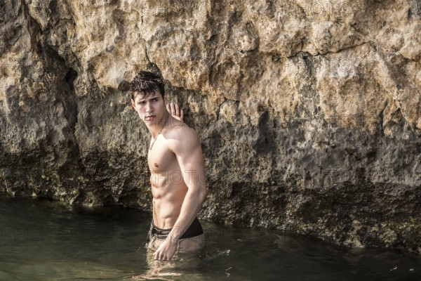 Attractive young shirtless athletic man standing in water by sea or ocean shore, wearing shorts, looking at camera, with rocks behind him