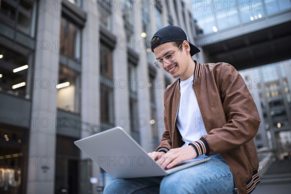 Happy handsome student looking at computer screen while typing something on laptop keyboard during work break outdoors, teenager looking at the notebook sitting at street