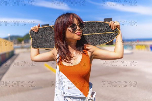 Portrait of a urban confident asian woman holding a skateboard on shoulders standing in an urban skateboard park under the sun in summer