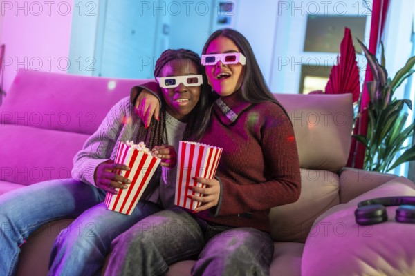 Two girls wearing 3d glasses enjoying a movie night at home, eating popcorn from striped boxes and having fun on the sofa