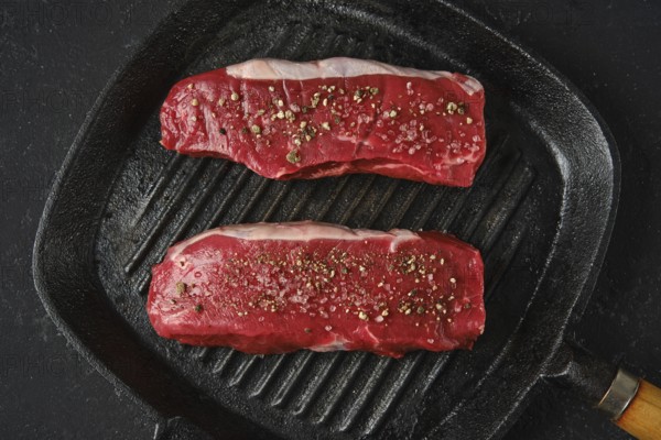 Top view of two thick cuts of raw beef steaks on a cast iron skillet. The marbled texture of the meat is seasoned with sea salt and pepper