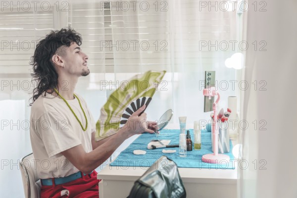 Young influencer with mullet hairstyle is cooling himself with a hand fan while applying makeup in front of a mirror in his bedroom, showing diversity and inclusion