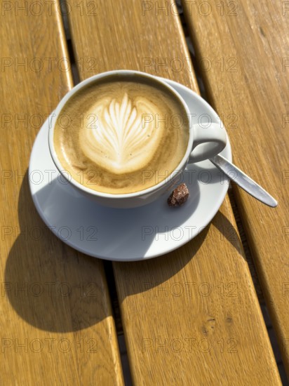 Coffee drink Cappuccino Cappuccio with flower pattern in milk foam, next to it single roasted candied almond on rustic table of outdoor café, international