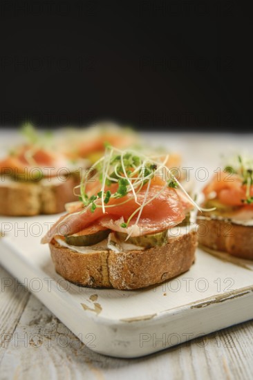 Open-faced sandwiches are arranged on a wooden platter, featuring layers of smoked salmon, pickles, and sprouts. This appetizing display highlights vibrant colors and fresh ingredients.