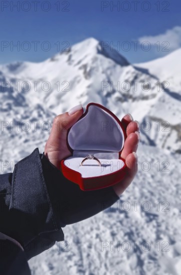 Female hand closeup holding an engagement ring inside a red heart shaped box over the snowy mountain peaks background. Marriage proposal on the mount top