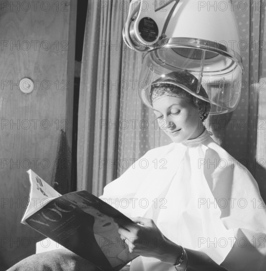 Hair dryer in the 1950s. A woman is sitting comfortably and reading Vogue magazine, while drying her hair under a drying hood. Sweden March 1954