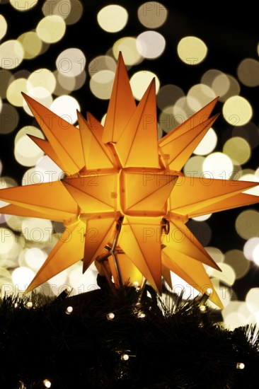 An orange-coloured, Christmassy star shines in the foreground in front of a beautiful, blurred bokeh background with lots of glittering, sparkling lights at a Christmas market at night, Cologne, North Rhine-Westphalia, Germany, Europe