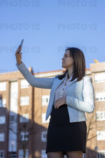 Business, middle-aged, brown-haired woman in a white shirt, black skirt and blue jacket is making selfies with her smartphone during her work time outdoors. Vertical portrait