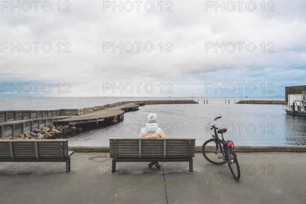 A young Caucasian woman sits with her back on a wooden bench overlooking the Baltic Sea on the seafront in Copenhagen Denmark in winter in cloudy weather. Girl walking gonoskoy bike parked nearby