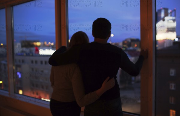 Back shot of young loving couple by the window at night. They hugging and enjoying city view. Romantic moments