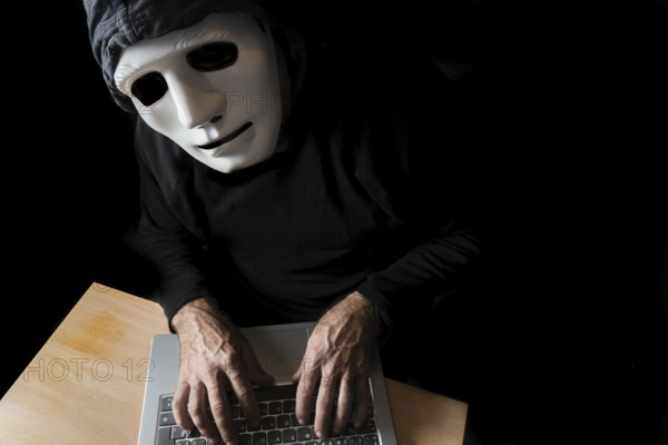 Hacker dressed in black with mask and hood typing on a computer while looking at the camera cybercrime concept